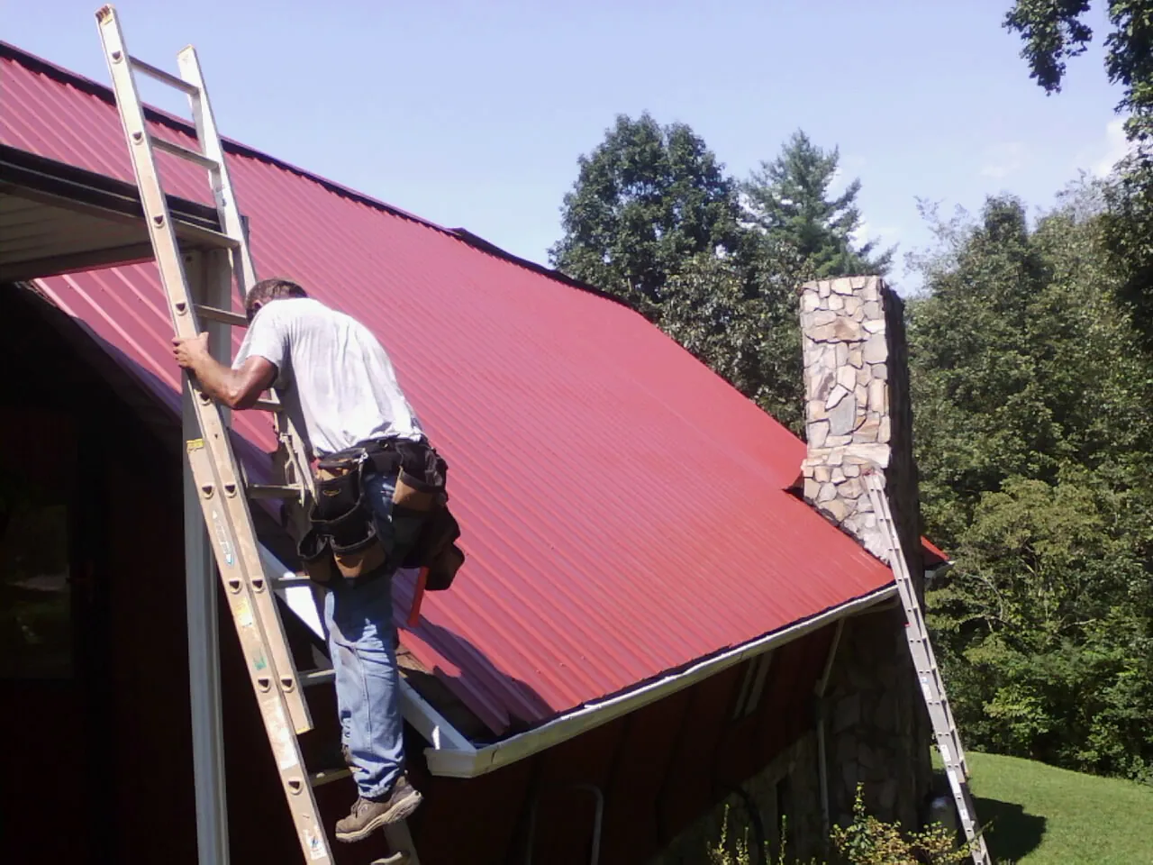 Roofer on a ladder beside a red metal roof and stone chimney — Soffit & Fascia Repair by Roofing Brothers in Franklin Furnace